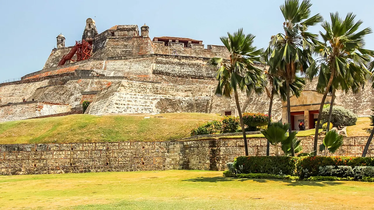 Muralhas e rampas de pedra do castelo de San Felipe de Barajas subindo um morro gramado, com guaritas no alto e palmeiras em primeiro plano.