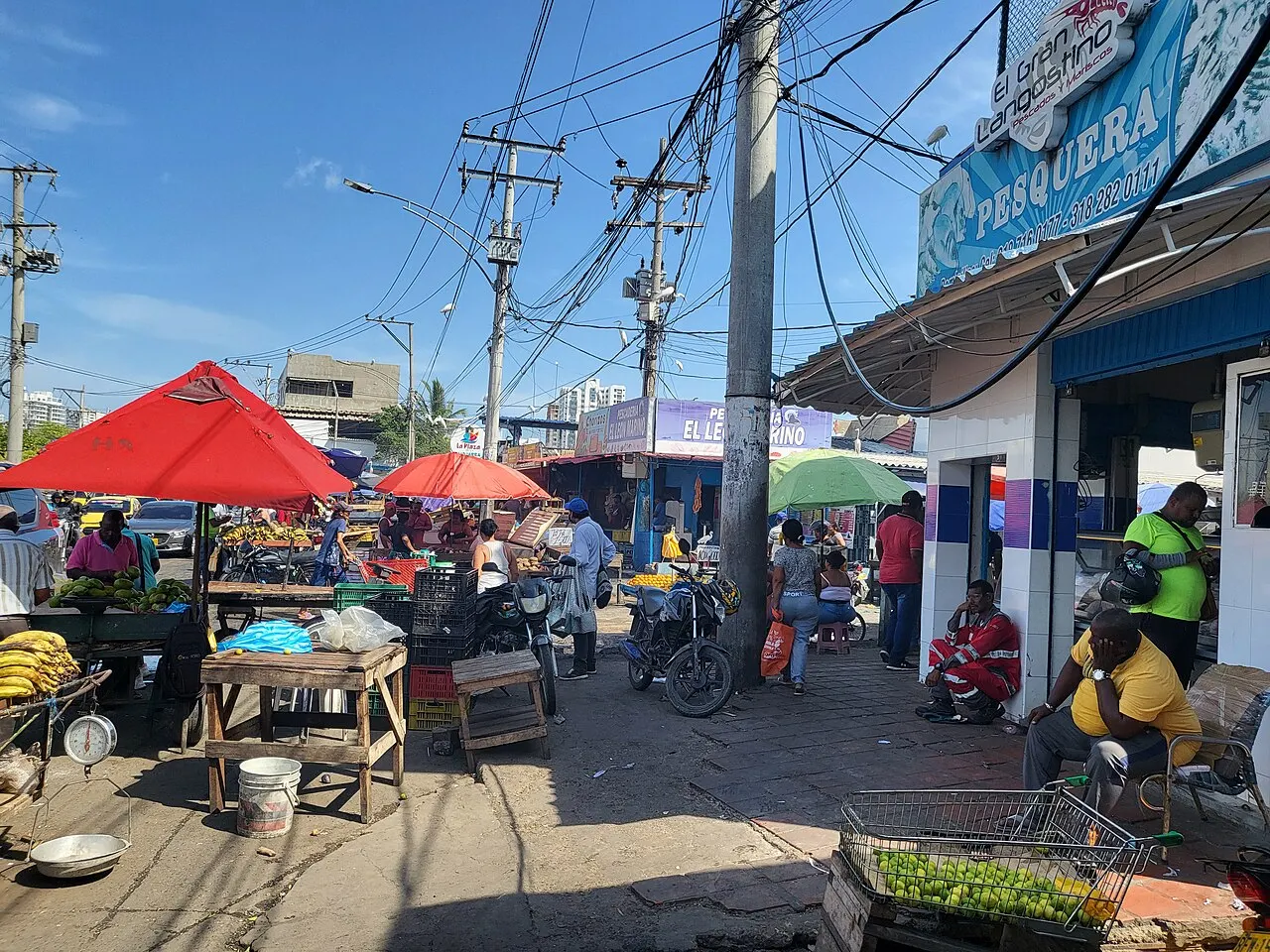Uma rua de mercado popular com barracas de frutas sob guarda-sóis vermelhos e verdes, caixas plásticas empilhadas, motos estacionadas e pessoas sentadas nas calçadas, com fios elétricos cruzando o céu azul.