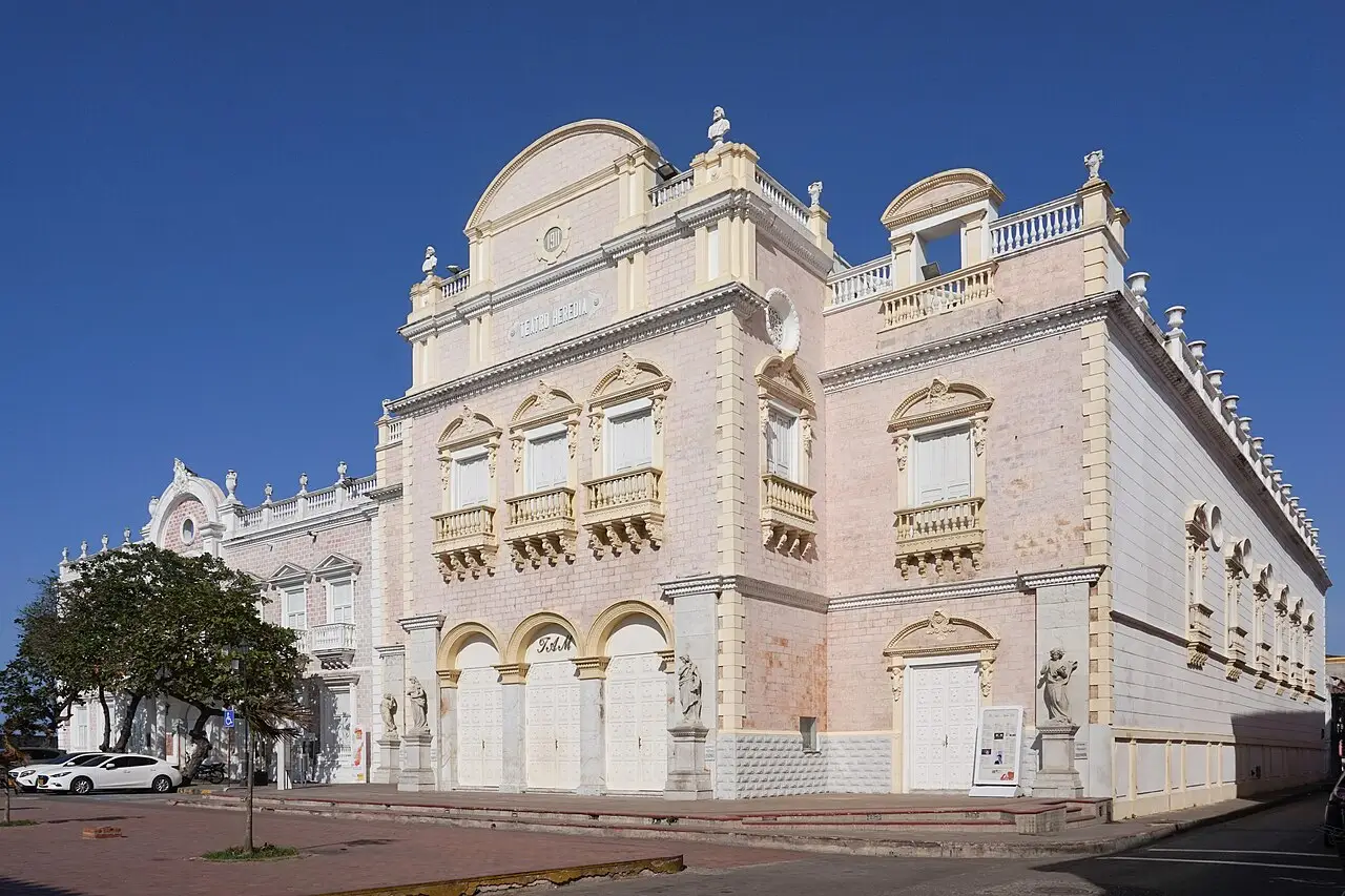 Fachada do Teatro Adolfo Mejía, de pedra clara, com janelas de moldura amarela, arcos no térreo e uma árvore na calçada.