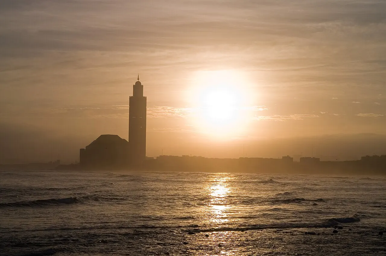 A silhueta escura de uma mesquita com um alto minarete retangular, à beira-mar, contra um sol brilhante nascendo entre nuvens, com ondas quebrando em primeiro plano.