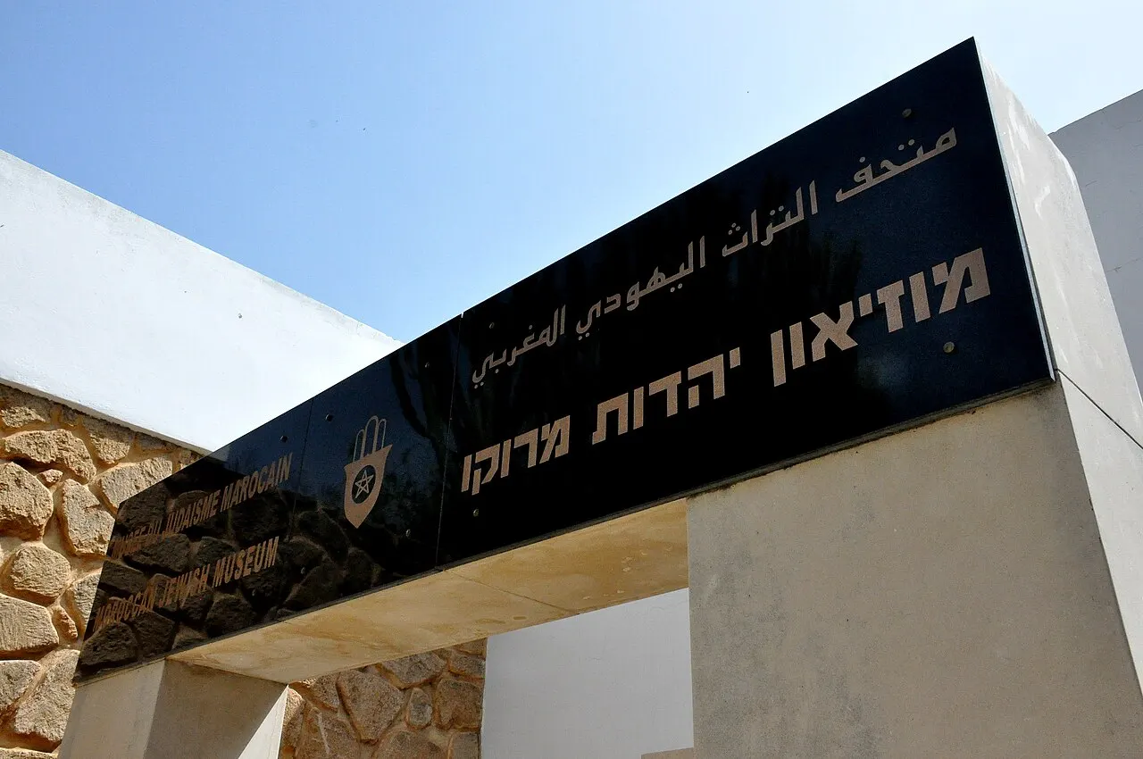 Placa preta sobre o pórtico do Museu do Judaísmo Marroquino, em Casablanca, com o nome em hebraico, árabe, francês e inglês e uma mão de Fátima gravada, sobre pilares de pedra e reboco branco.