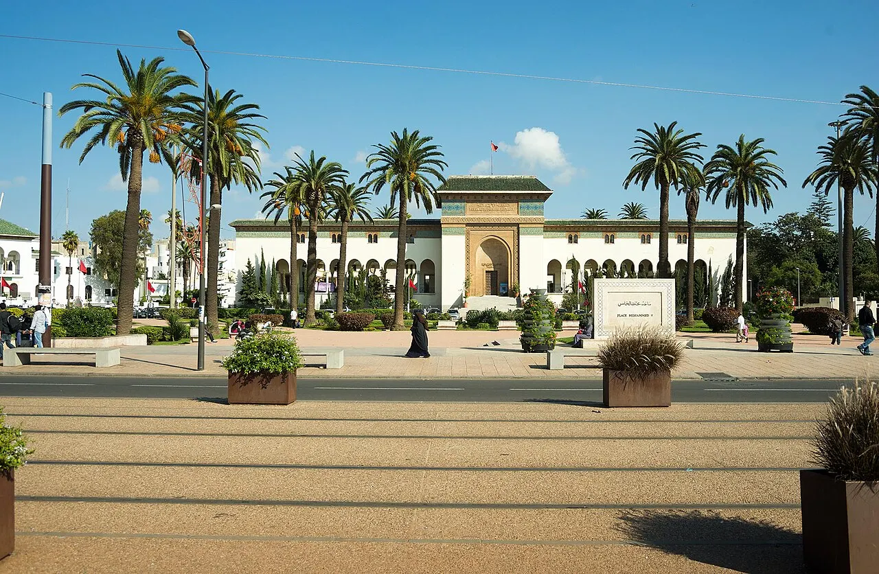 A fachada branca de um prédio institucional em estilo mourisco, com um grande portal ornamentado e telhado verde, cercada de palmeiras altas e jardins com canteiros, sob céu azul.