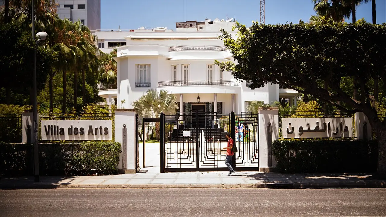 Villa des Arts, casa branca de dois andares atrás do portão, com o letreiro do nome e árvores na frente.