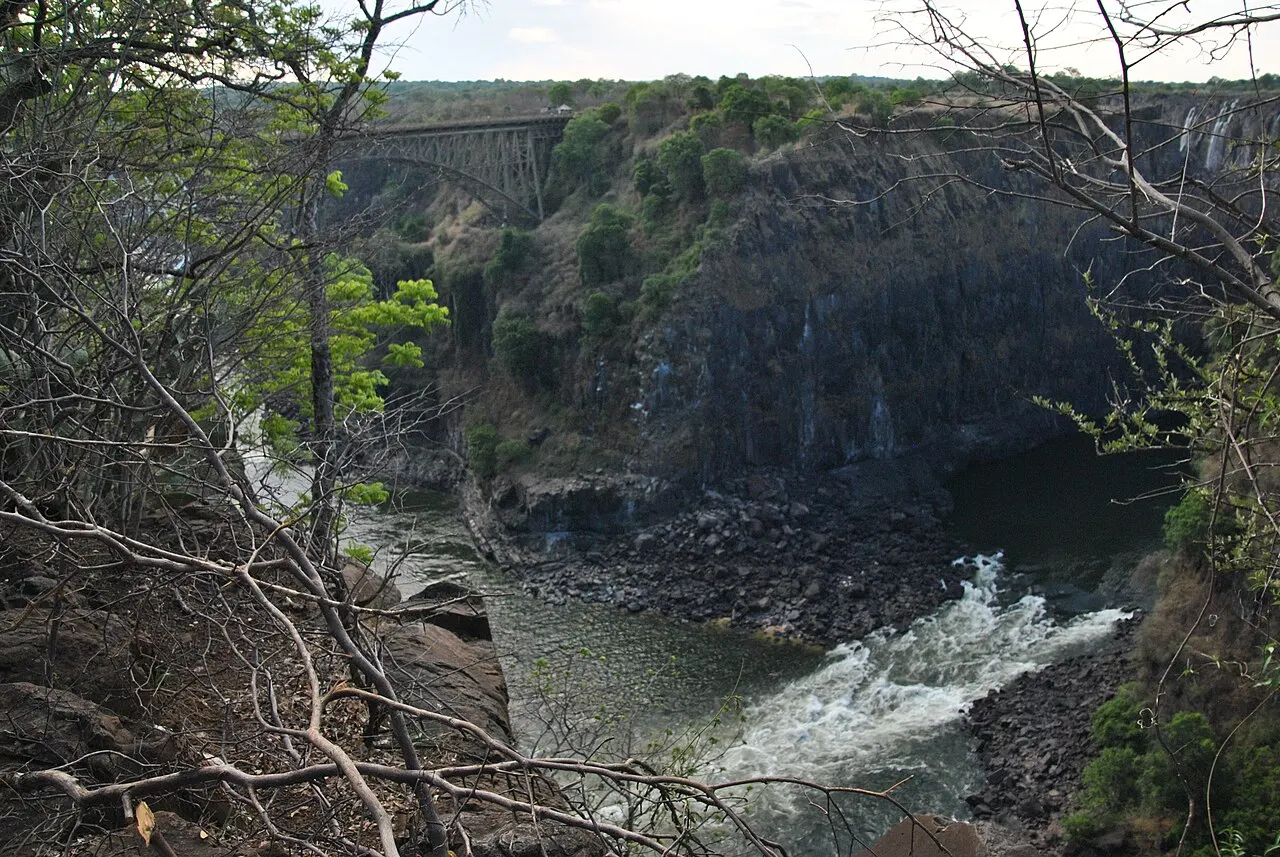La rivière descend en rapides blancs au fond d'une gorge aux parois de roche sombre, vue entre des branches sèches, avec un pont d'acier au loin.
