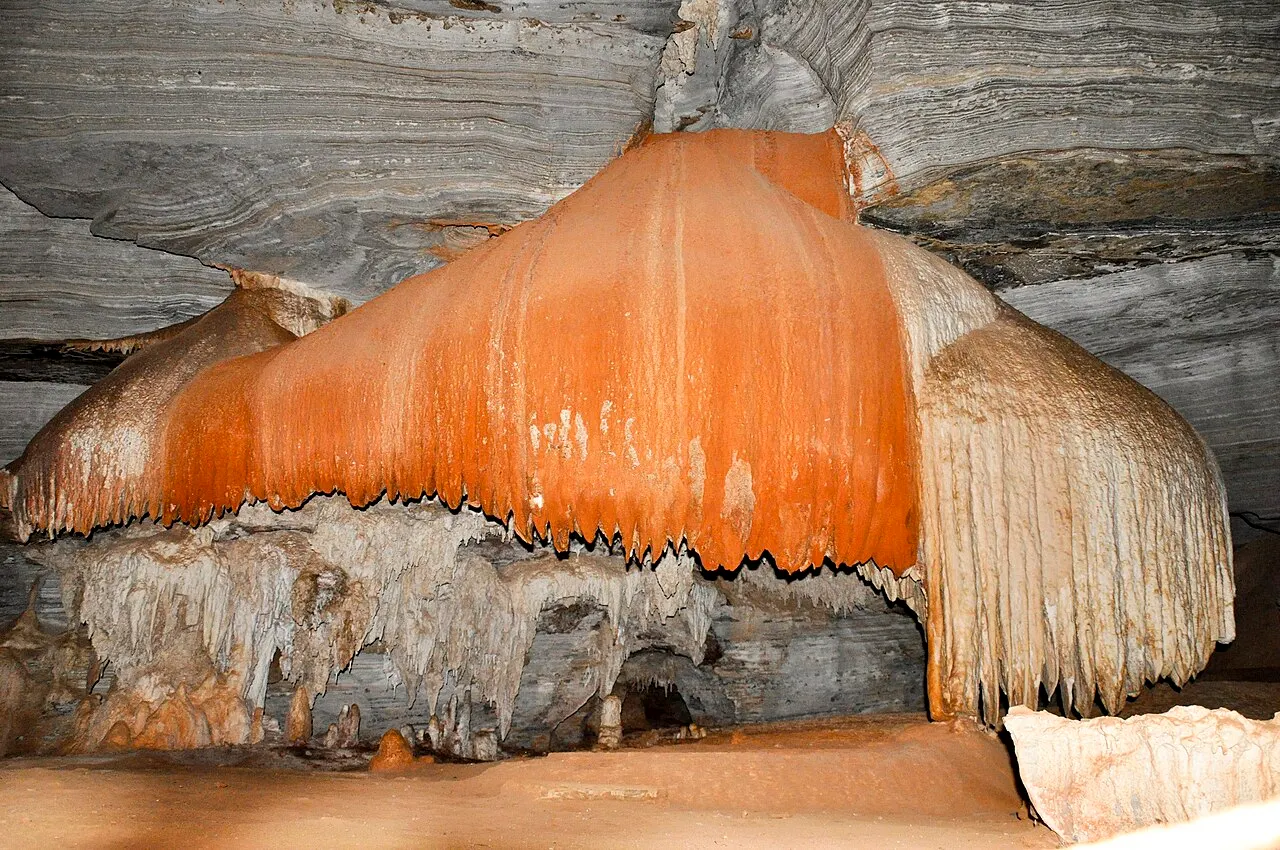 Uma grande formação de calcário alaranjada, como uma cortina escorrida, dentro do salão da Gruta da Lapa Doce.