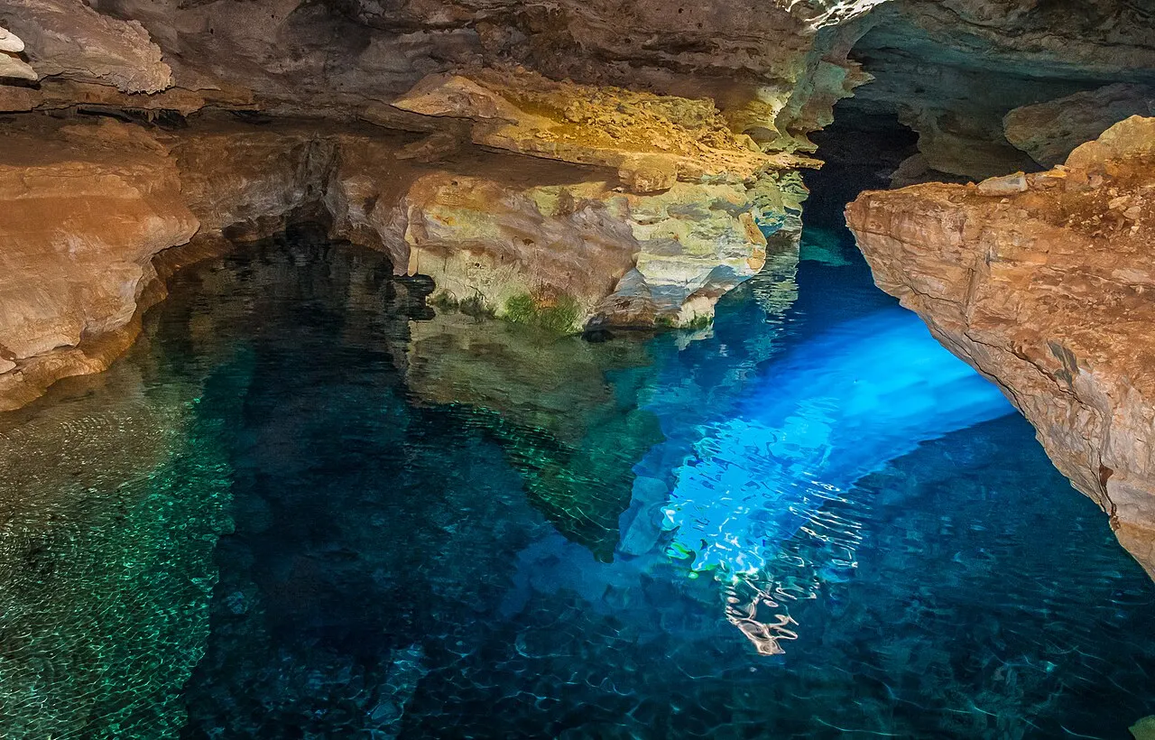 Um feixe de sol entrando pela gruta e acendendo de azul a água transparente do Poço Azul, entre rochas coloridas.