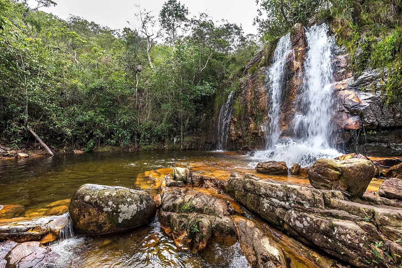 Uma queda da Cachoeira dos Cristais descendo entre a mata até um poço raso com pedras na frente.