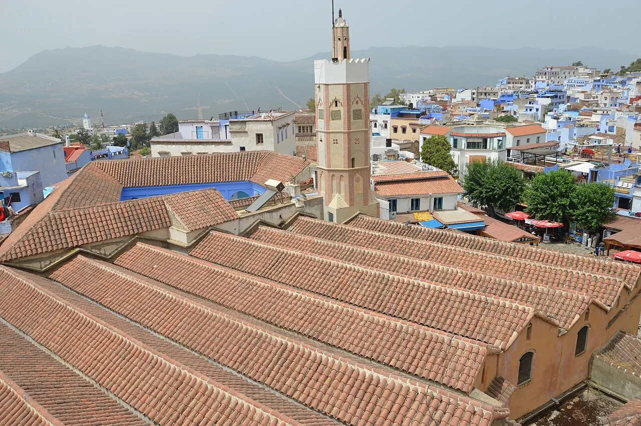 Haut minaret au-dessus de longs toits de tuiles rouges ; autour, les maisons blanches et bleues de la médina et, au loin, des montagnes dans la brume.