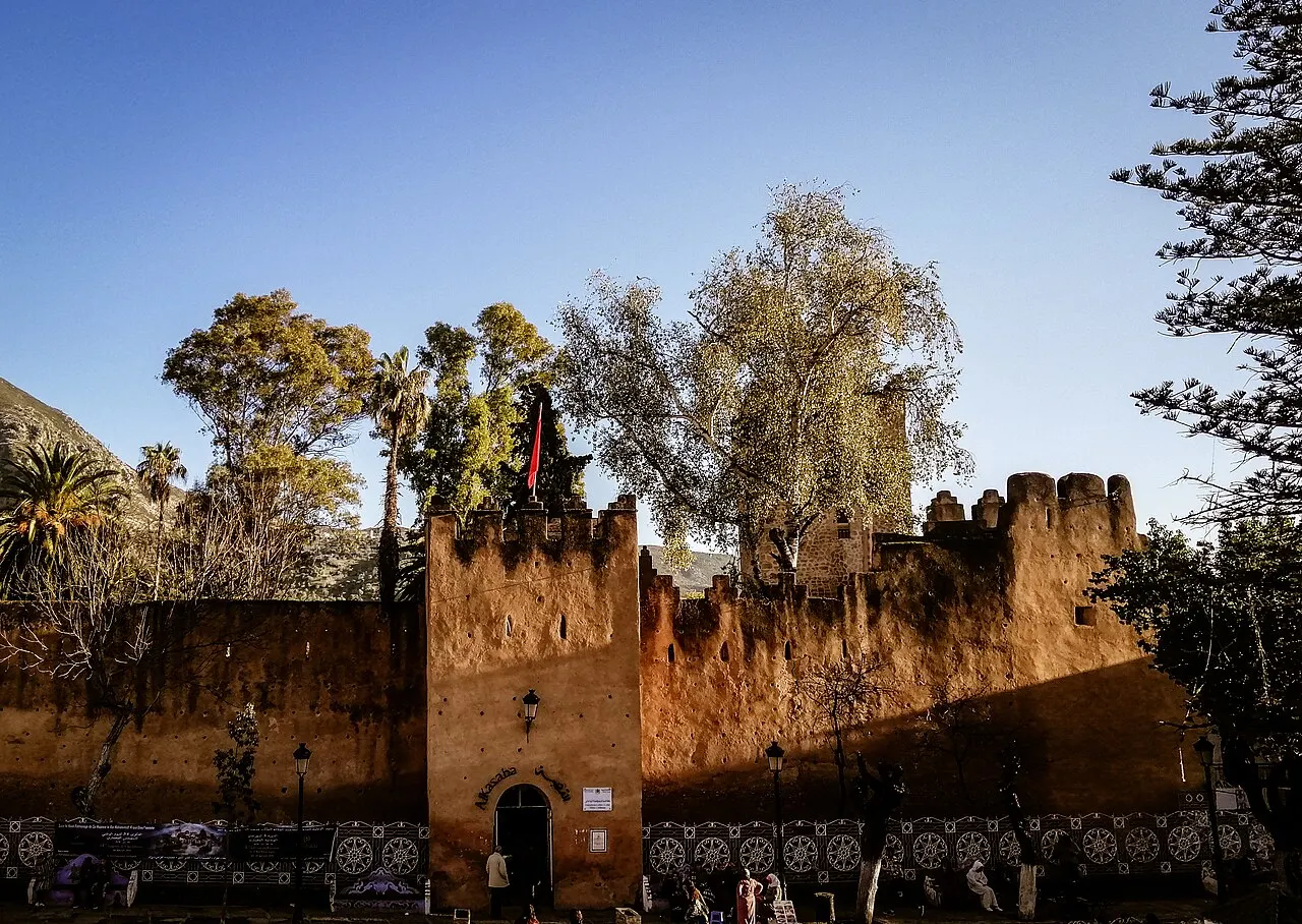 Rempart crénelé en pisé rougeâtre avec une tour carrée et la porte d'entrée en arc, dans la lumière de fin d'après-midi ; de grands arbres dépassent derrière les créneaux.