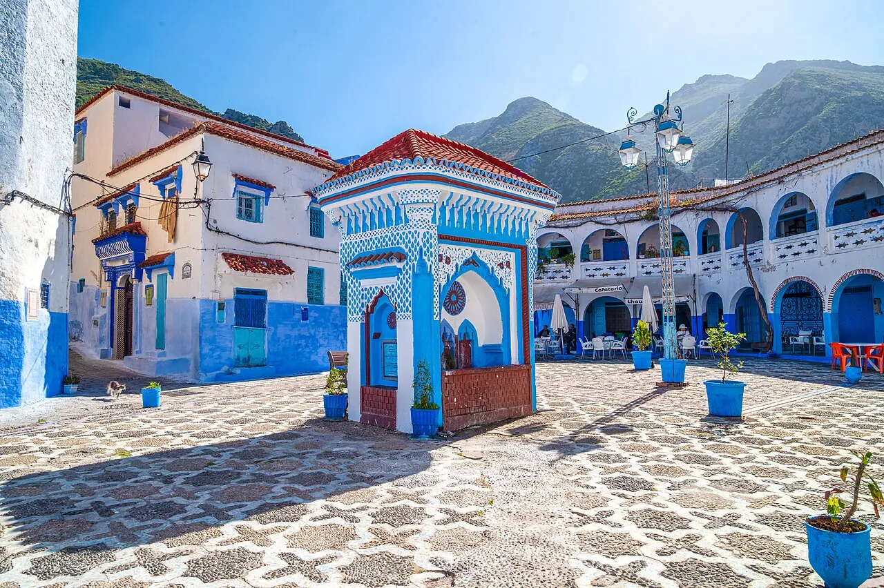 Place pavée avec une fontaine en zellige bleu et blanc sous un toit de tuiles rouges, des pots bleus, des maisons blanches et bleues et une galerie à arcades ; montagnes au fond sous un ciel dégagé.