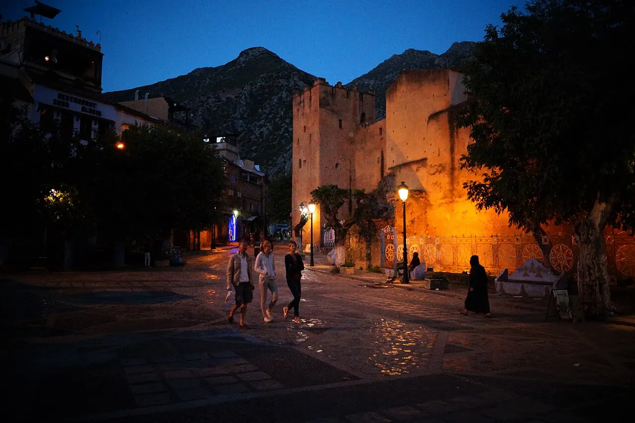 Place pavée à la tombée de la nuit, avec des passants et des réverbères allumés devant un rempart en pisé éclairé ; derrière, le versant de la montagne sous un ciel bleu sombre.