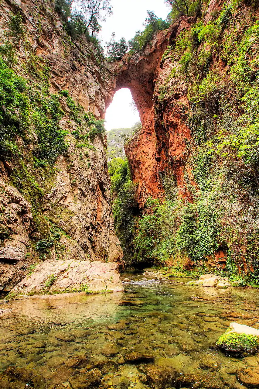 Arche naturelle de roche rougeâtre reliant les deux parois d'une gorge couverte de végétation, vue depuis le lit d'une rivière peu profonde aux eaux claires et caillouteuses.
