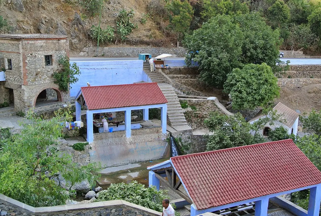 Deux lavoirs aux toits de tuiles rouges et aux piliers bleus au-dessus de bassins, à côté d'un bâtiment en pierre à arcades et d'un escalier qui monte le versant boisé.