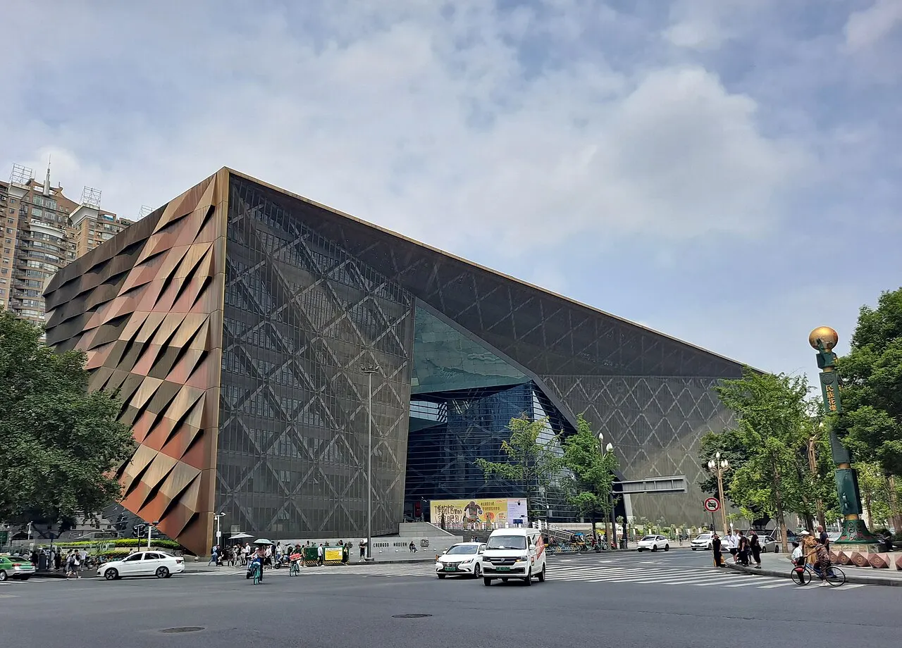 An angular building clad in diamond-patterned metal panels, with a copper-coloured facade and a green glass recess, on a street corner with cars and a cloudy sky.