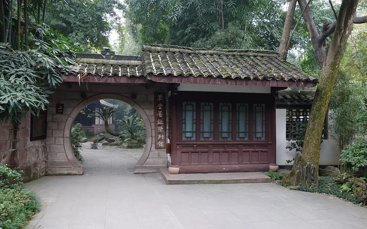 A small wooden pavilion with a tiled roof and a round doorway in the wall beside it, among bamboo and trees, with a stone path in front.