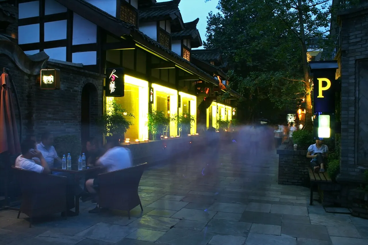 A stone alley at dusk with timber-framed white houses, shop windows lit in yellow, tables and armchairs outside and passers-by blurred by motion.
