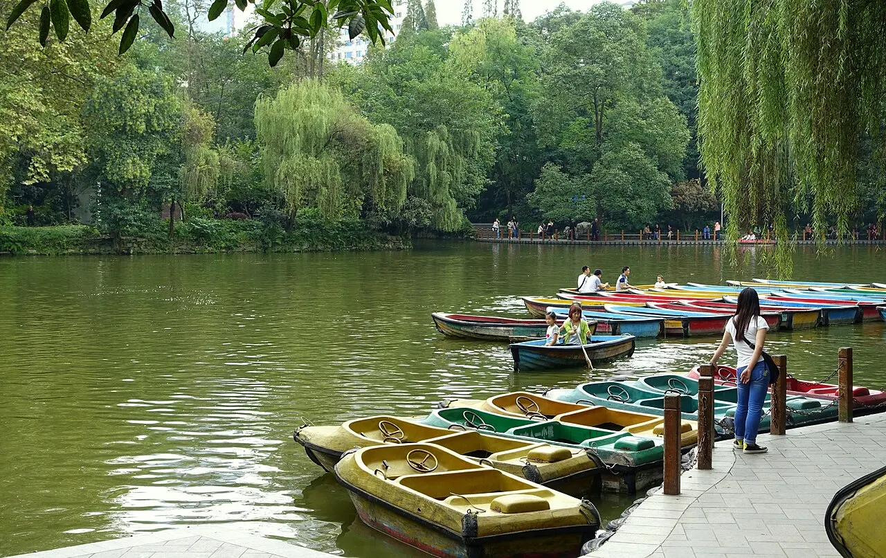 A green lake with colourful rowing boats moored at a wooden pier, willows and dense trees on the far bank.