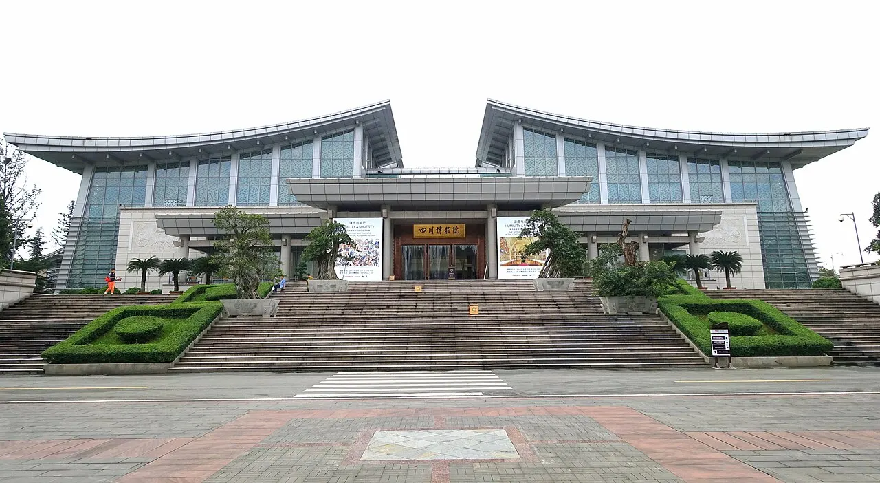 A glass and concrete building with two curved roofs rising at the ends, a wide staircase flanked by hedge beds and a paved plaza in front.
