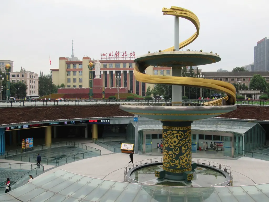 A column with a golden spiral and stacked discs in the middle of a sunken plaza with a pool; behind it, a pale-fronted building with a white statue in front.