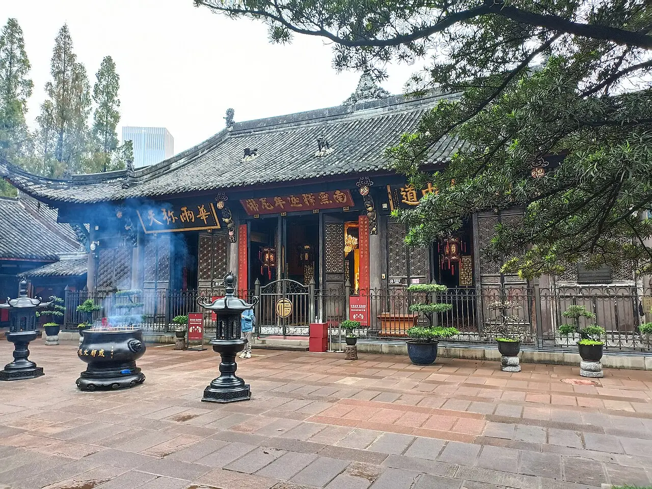 A wooden hall with a grey tiled roof and gilded boards over the entrance; in the paved courtyard a large incense burner gives off smoke between iron lanterns.