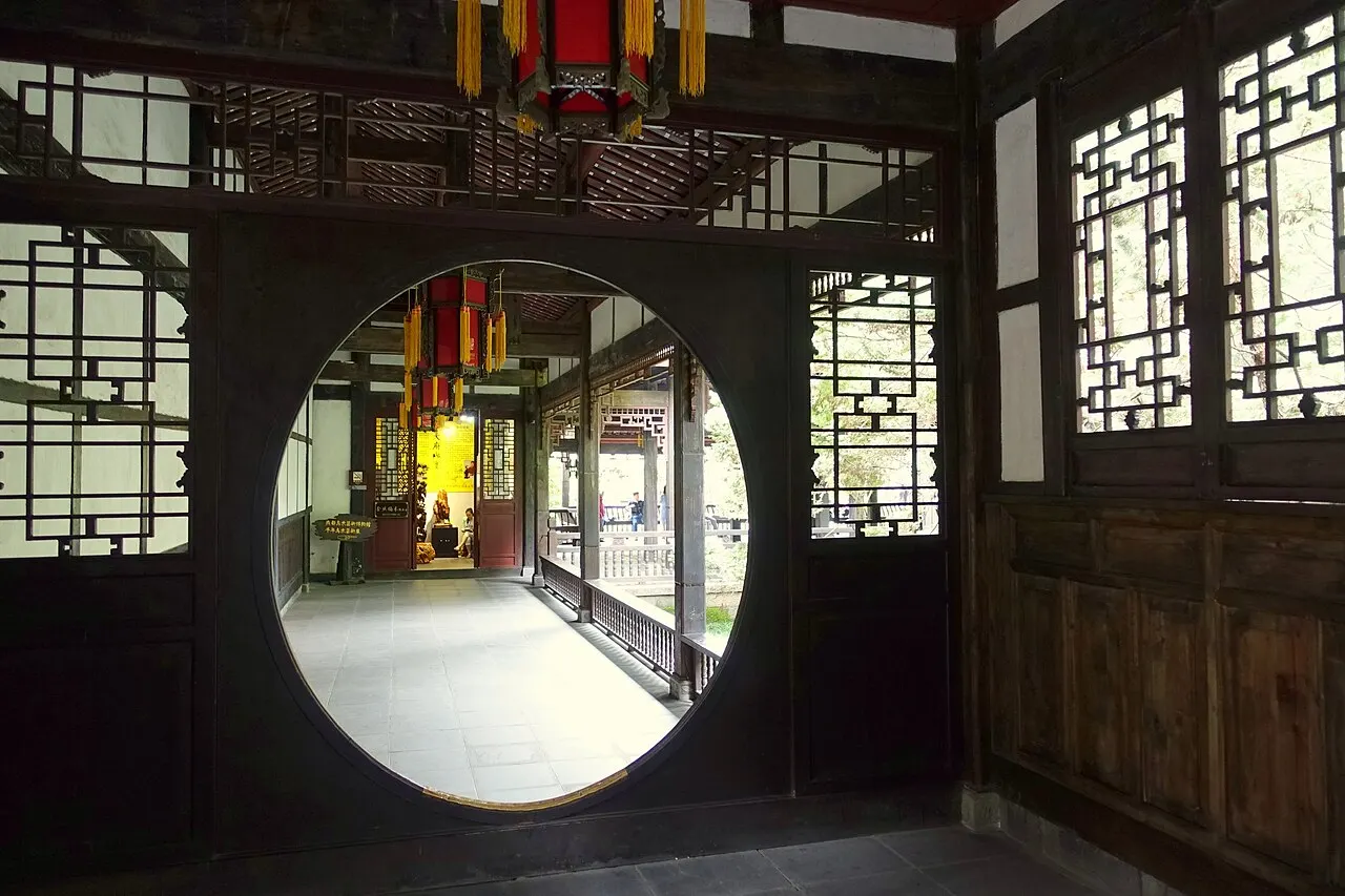 A dark wooden interior with lattice windows and a moon-shaped doorway opening onto a covered gallery, with lanterns hanging from the ceiling.