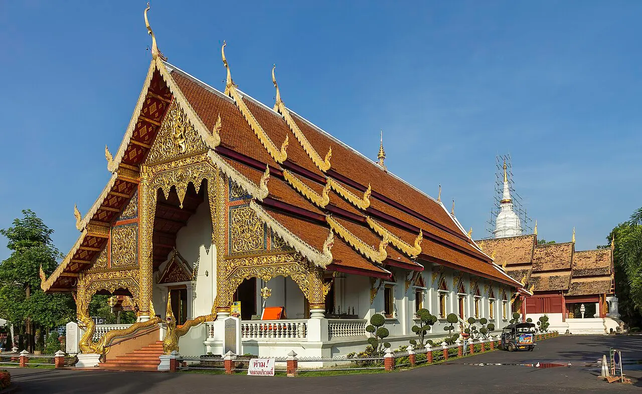 Templo tailandês de telhados vermelhos sobrepostos e frontão dourado, com um chedi branco atrás.