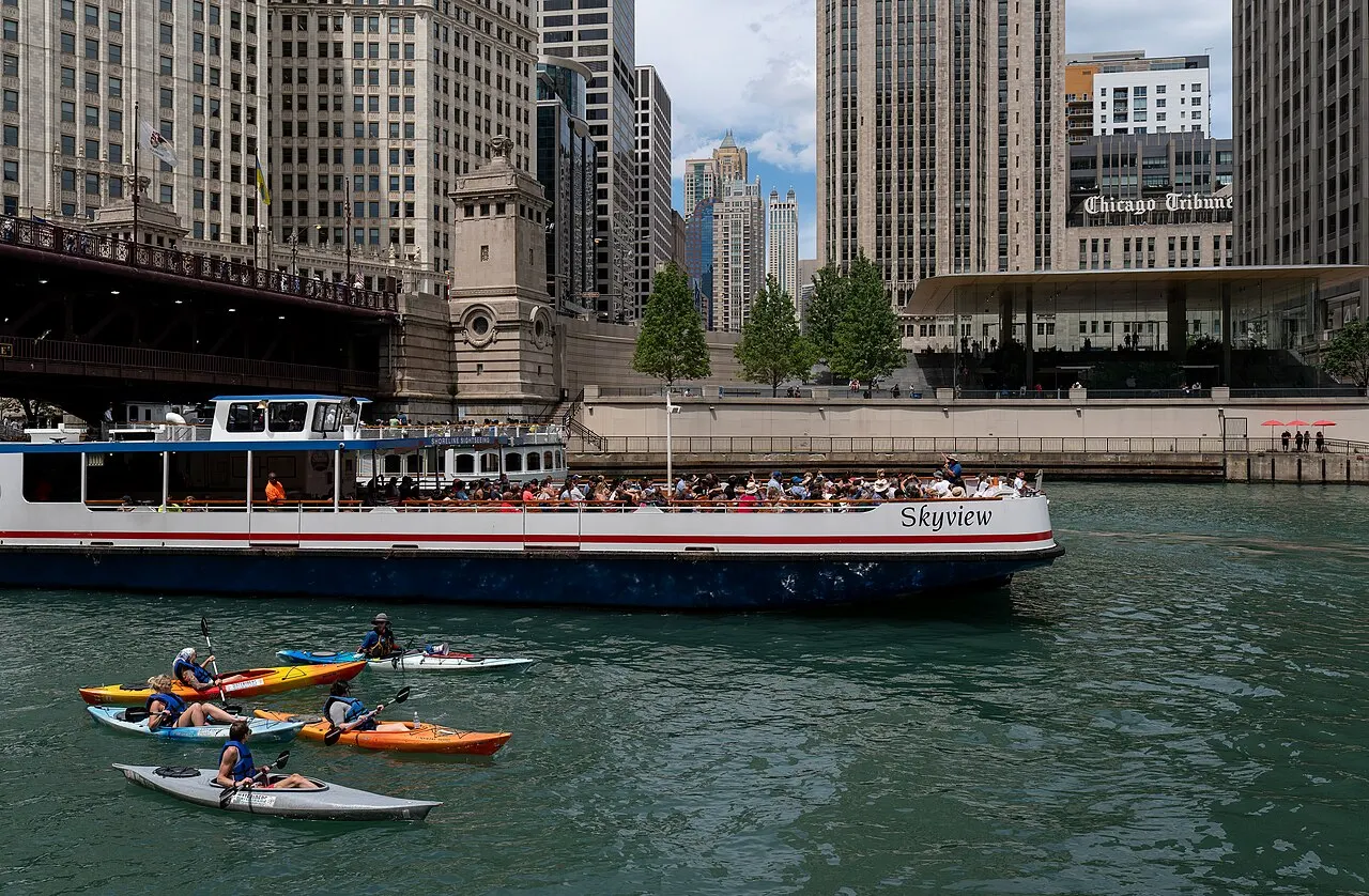 Barco de turismo lotado de passageiros navega o rio cercado de arranha-céus, com caiaques coloridos ao redor e uma ponte histórica ao fundo em dia nublado.