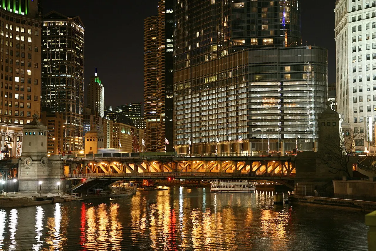 Rio de Chicago à noite, com uma ponte de aço iluminada, um barco branco e torres acesas refletidas na água.