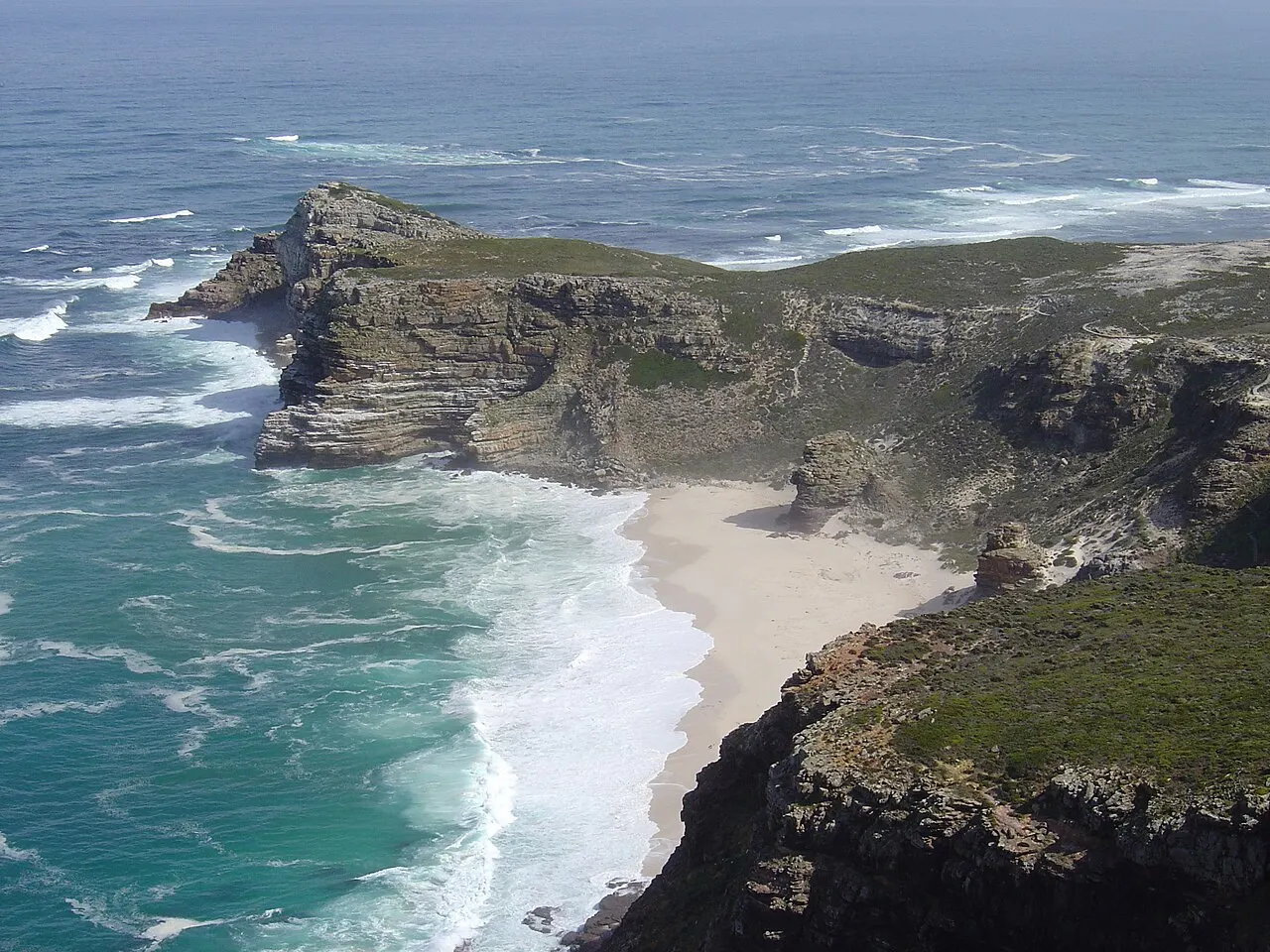 O costão do Cabo da Boa Esperança desce até a praia da enseada, entre o penhasco e o mar azul e verde.