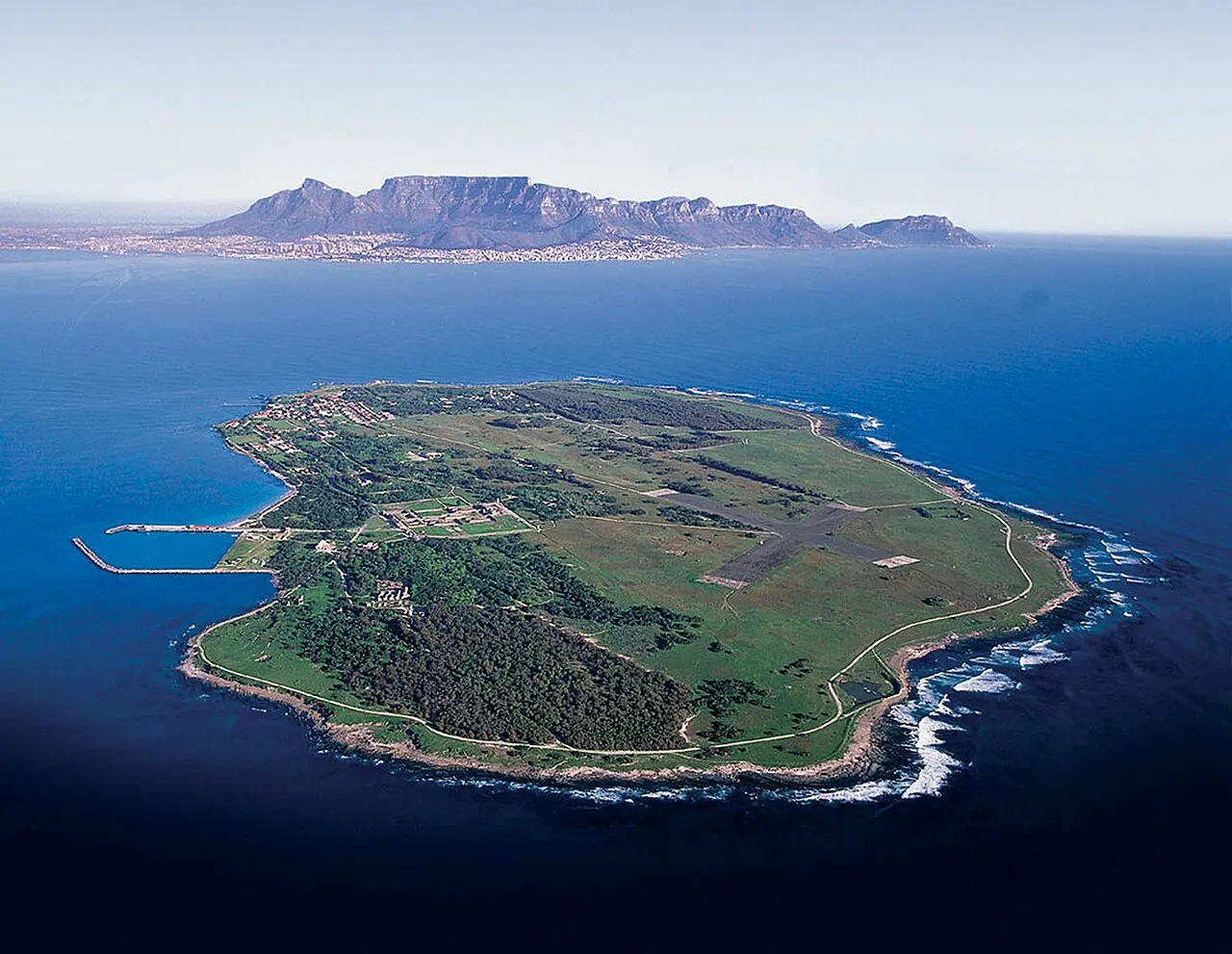Ilha de Robben, plana e verde, cercada pelo mar azul, vista do alto, com a costa ao fundo.