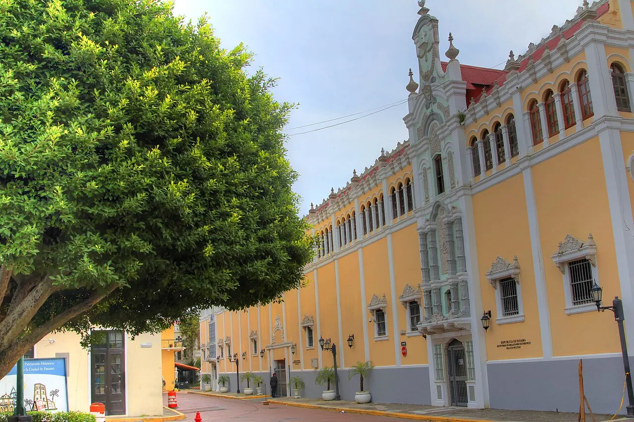 A fachada de um prédio colonial em amarelo, com molduras brancas ornamentadas, arcadas no andar de baixo e telhado vermelho, numa rua sombreada por uma grande árvore, sob céu claro.