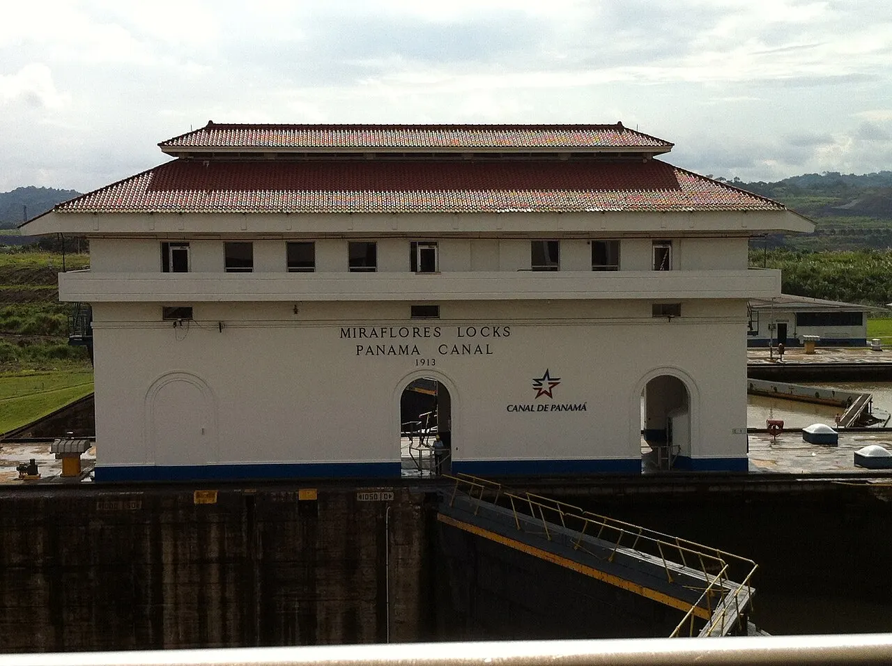 Prédio principal das eclusas de Miraflores, no canal do Panamá: um edifício comprido e baixo, de paredes claras e telhado escuro de duas águas, com um brasão no centro da fachada.