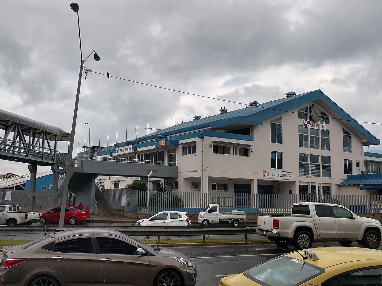 Prédio branco de telhado azul do Mercado de Mariscos da Cidade do Panamá, com o nome na fachada e as bandeiras do Panamá e do Japão no frontão, uma passarela de pedestres à esquerda e carros e um táxi amarelo na avenida em primeiro plano, sob céu carregado.