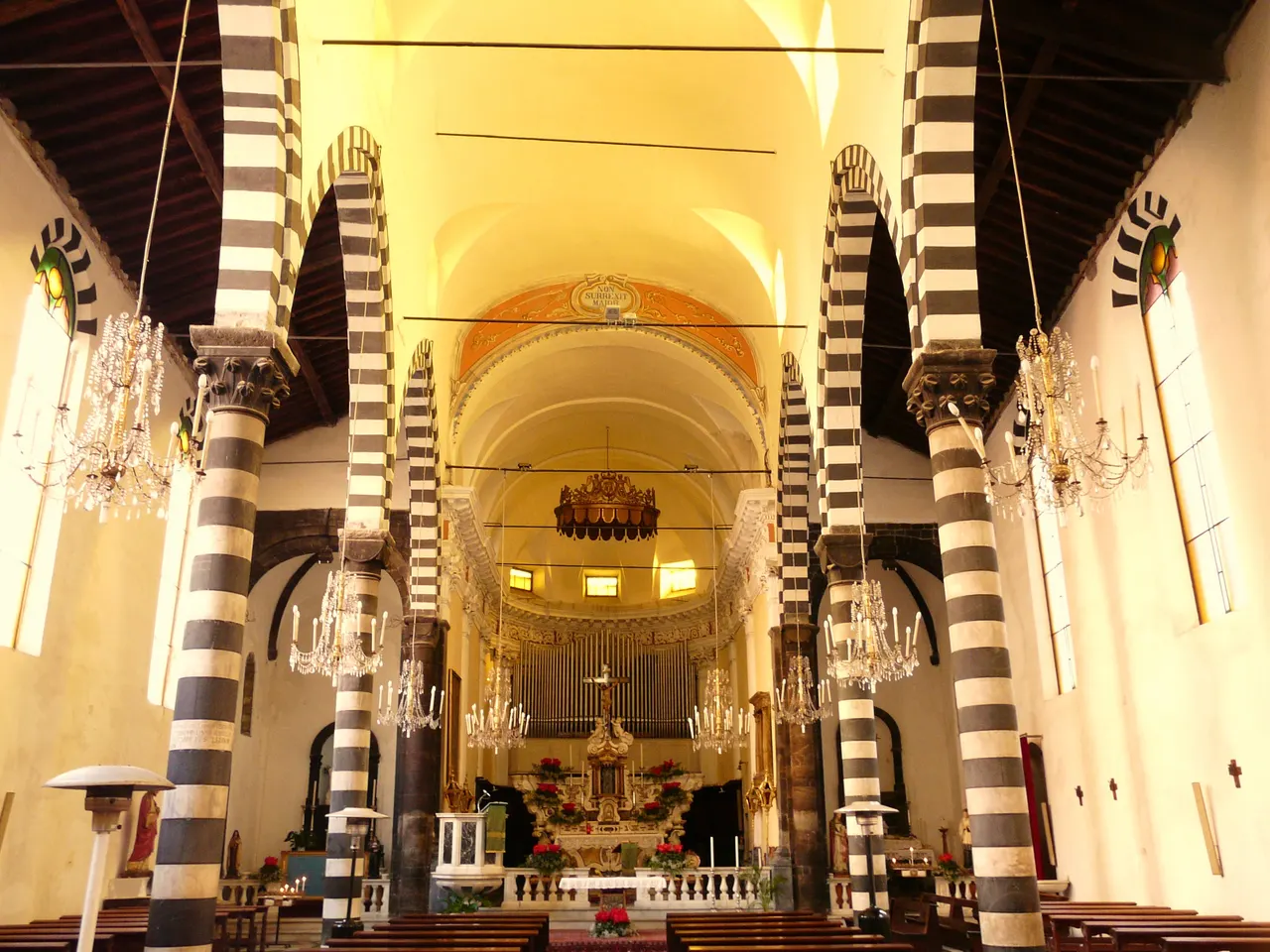 A nave de San Giovanni Battista, em Monterosso al Mare, com colunas e arcos de faixas claras e escuras, lustres, bancos de madeira e o altar ao fundo.