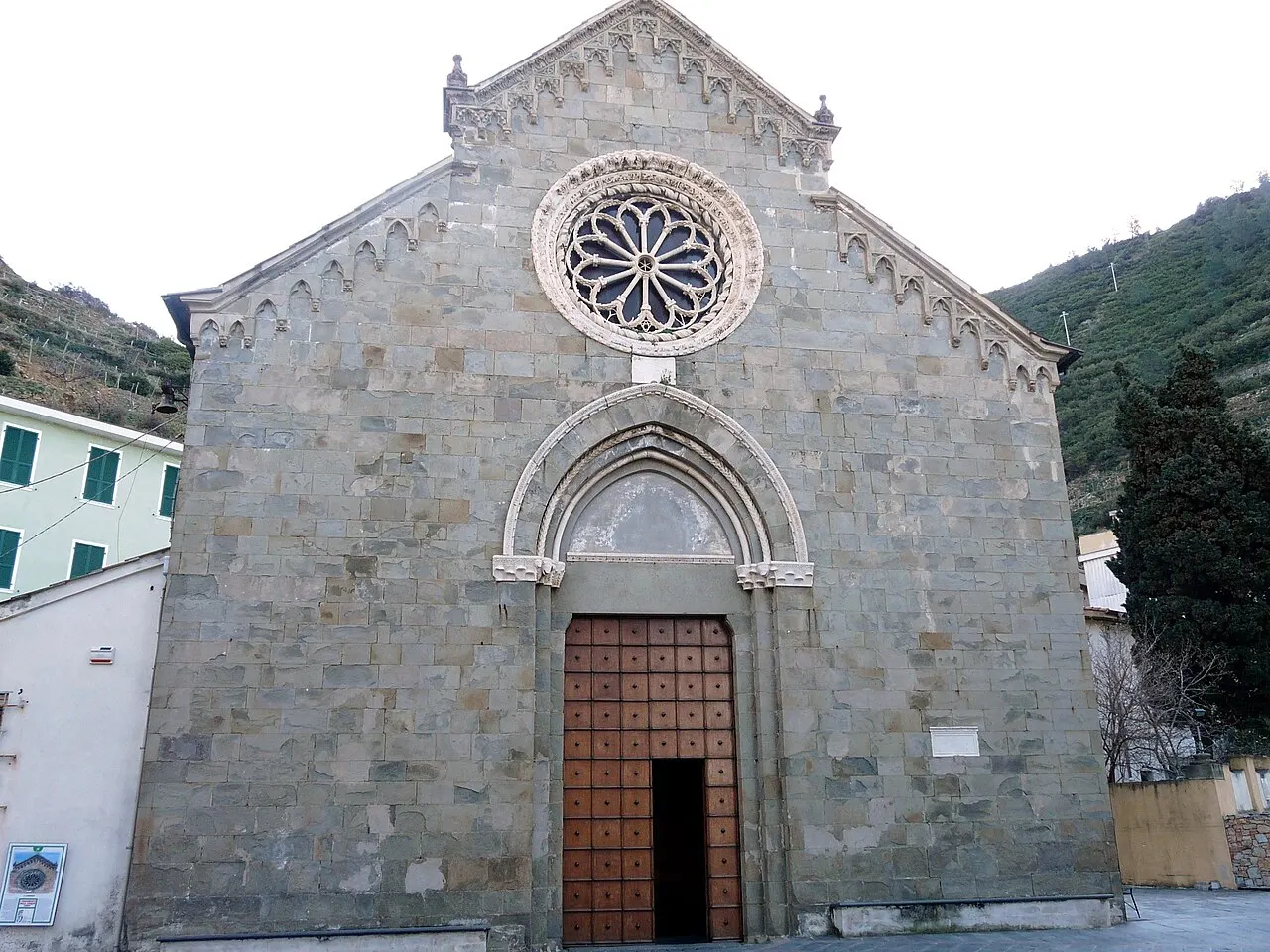 A fachada de pedra cinza da igreja de San Lorenzo, em Manarola, com uma rosácea de mármore branco sobre a porta de madeira.