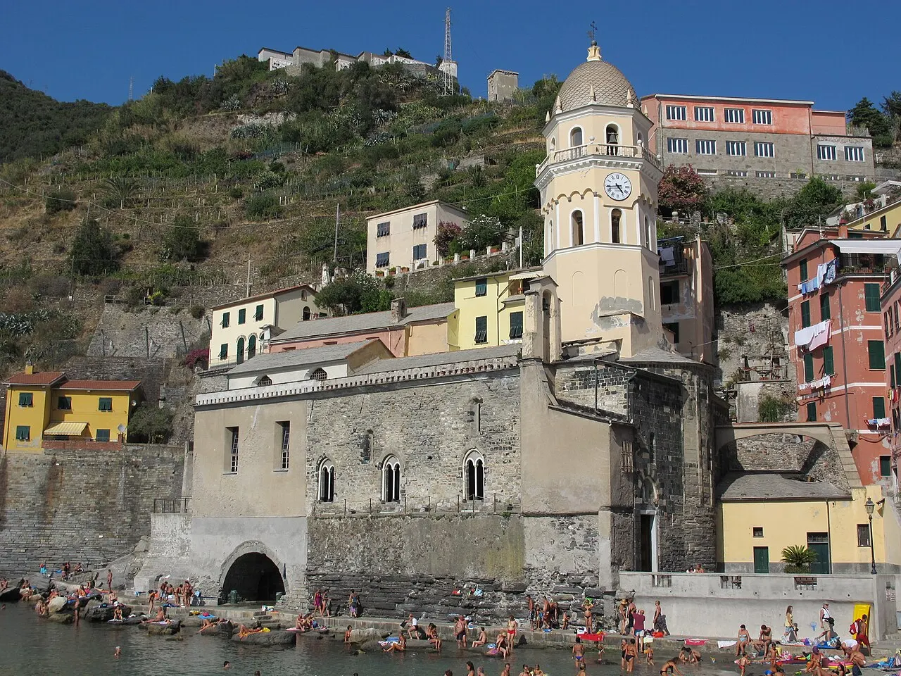 A igreja de Santa Margherita di Antiochia na beira do porto de Vernazza, com a torre octogonal do sino e banhistas nas pedras em frente.