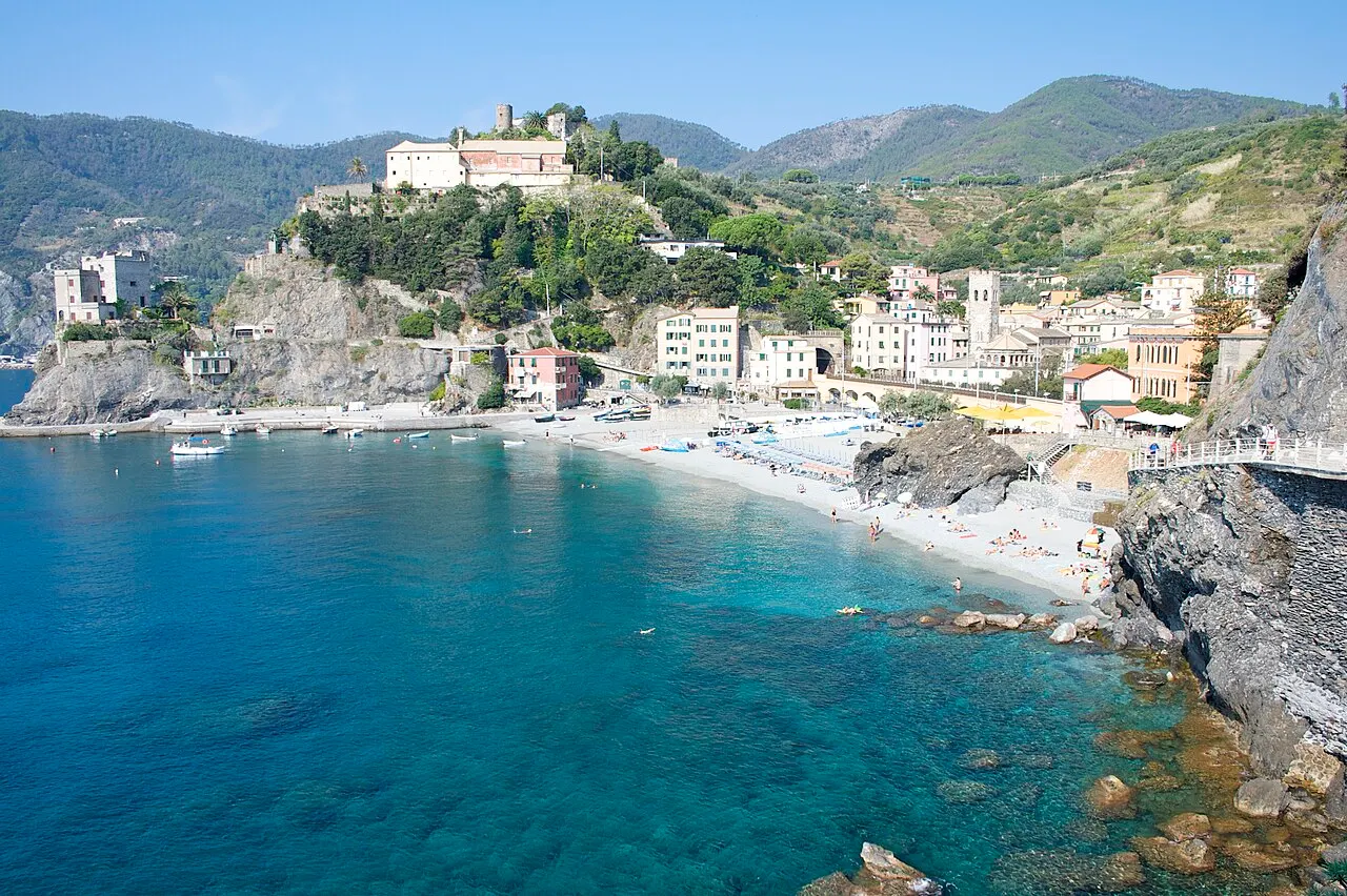 A baía de Monterosso com água verde-azulada, a praia e a vila; no morro acima das casas, o convento dos capuchinhos entre árvores.