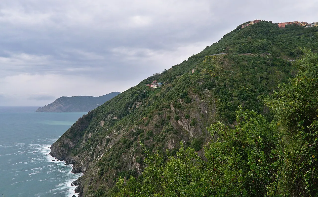 A encosta verde e íngreme que o Sentiero Azzurro atravessa perto de Manarola, caindo no mar, com um cabo ao fundo sob céu nublado.