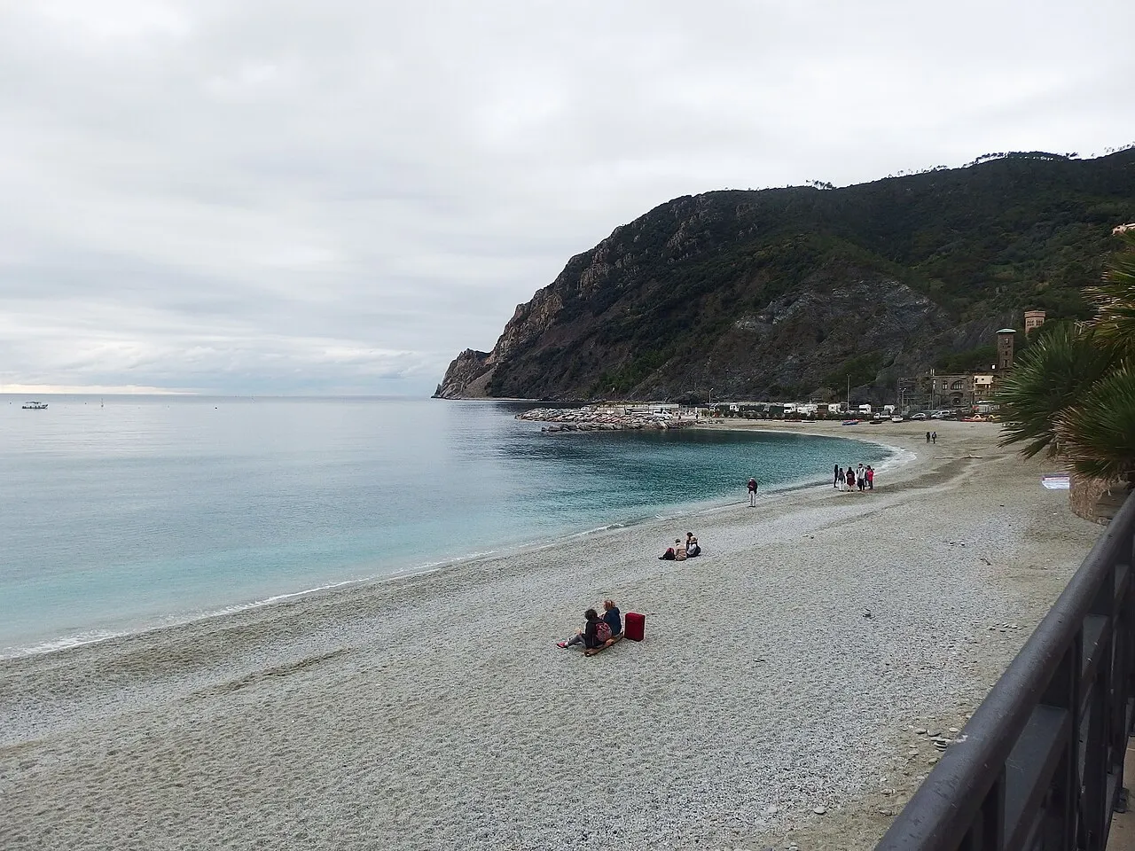 A praia de seixos de Fegina, em Monterosso, quase vazia num dia nublado, com o mar calmo e o cabo de Punta Mesco ao fundo.