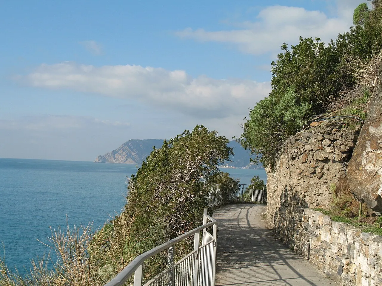 O caminho pavimentado da Via dell'Amore, com guarda-corpo, cortado na encosta de pedra sobre o mar, com vegetação e um cabo ao fundo.