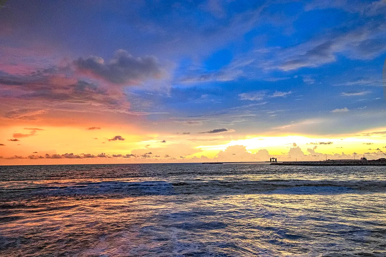 O mar visto da orla, com ondas quebrando em primeiro plano e o céu tingido de laranja, rosa e azul pelo pôr do sol, uma estrutura escura ao longe no horizonte.