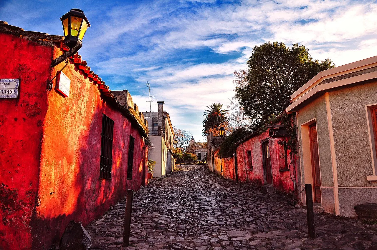 A Calle de los Suspiros, em Colonia del Sacramento, com calçamento de pedras irregulares descendo entre casas baixas vermelhas e rosadas, um lampião na parede e céu com nuvens.