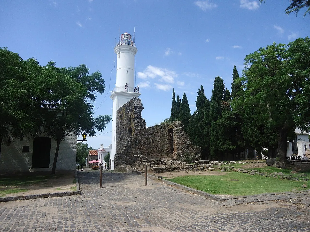 O farol branco de Colonia del Sacramento, alto e cilíndrico, ao lado das ruínas de pedra do convento de São Francisco, com ciprestes e calçamento de pedra.