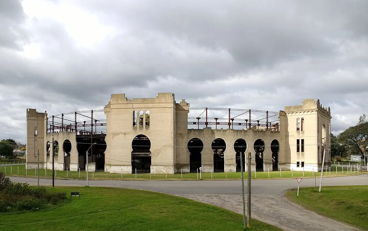 A arena de touros do Real de San Carlos, em Colonia del Sacramento, com fachada de arcos, torres nos cantos e estrutura metálica no alto, sob céu nublado, com gramado na frente.