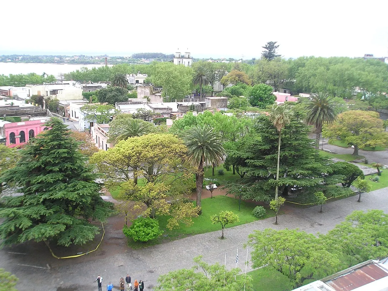 A Plaza Mayor de Colonia del Sacramento vista do alto, com árvores grandes, palmeiras e caminhos, e as casas baixas do Barrio Histórico em volta.