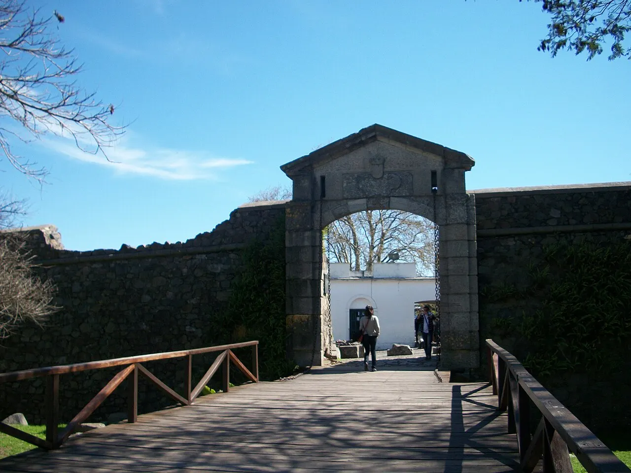 O Portón de Campo, em Colonia del Sacramento, com o arco de pedra na muralha, a ponte de madeira sobre o fosso e pessoas passando, sob céu azul.