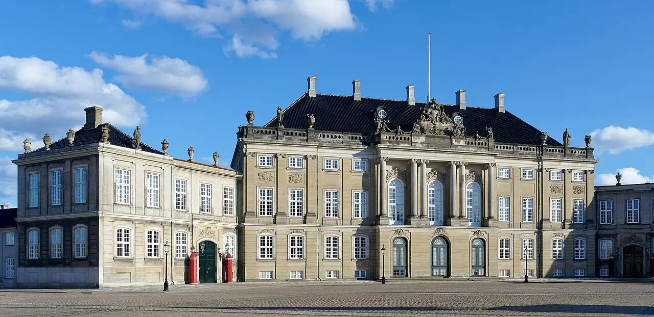 Fachada do palácio de Amalienborg na praça, com telhado escuro, estátuas no alto e duas guaritas vermelhas.