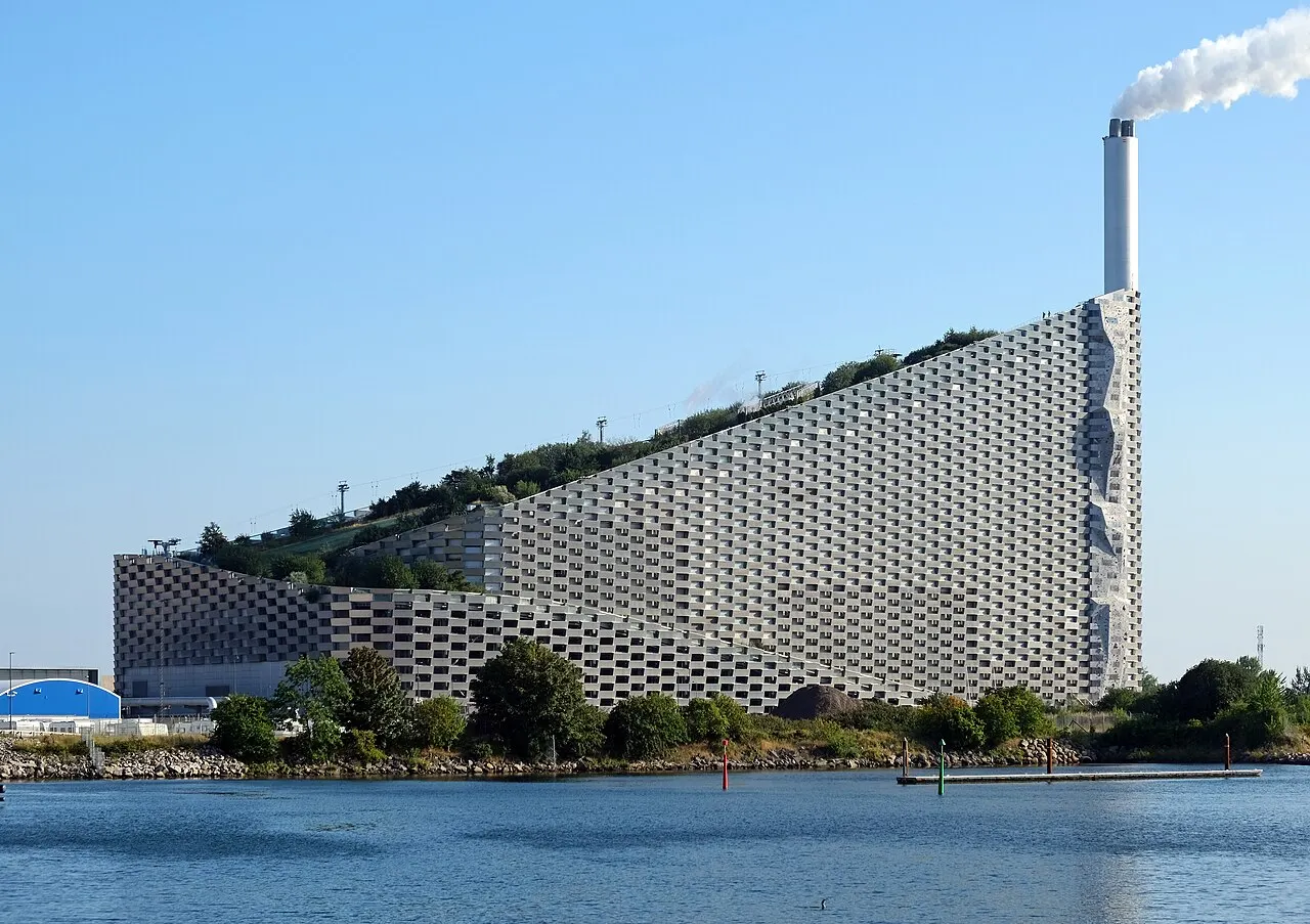 O CopenHill aparece da água, com a fachada inclinada, a rampa no telhado e uma coluna branca saindo da chaminé.