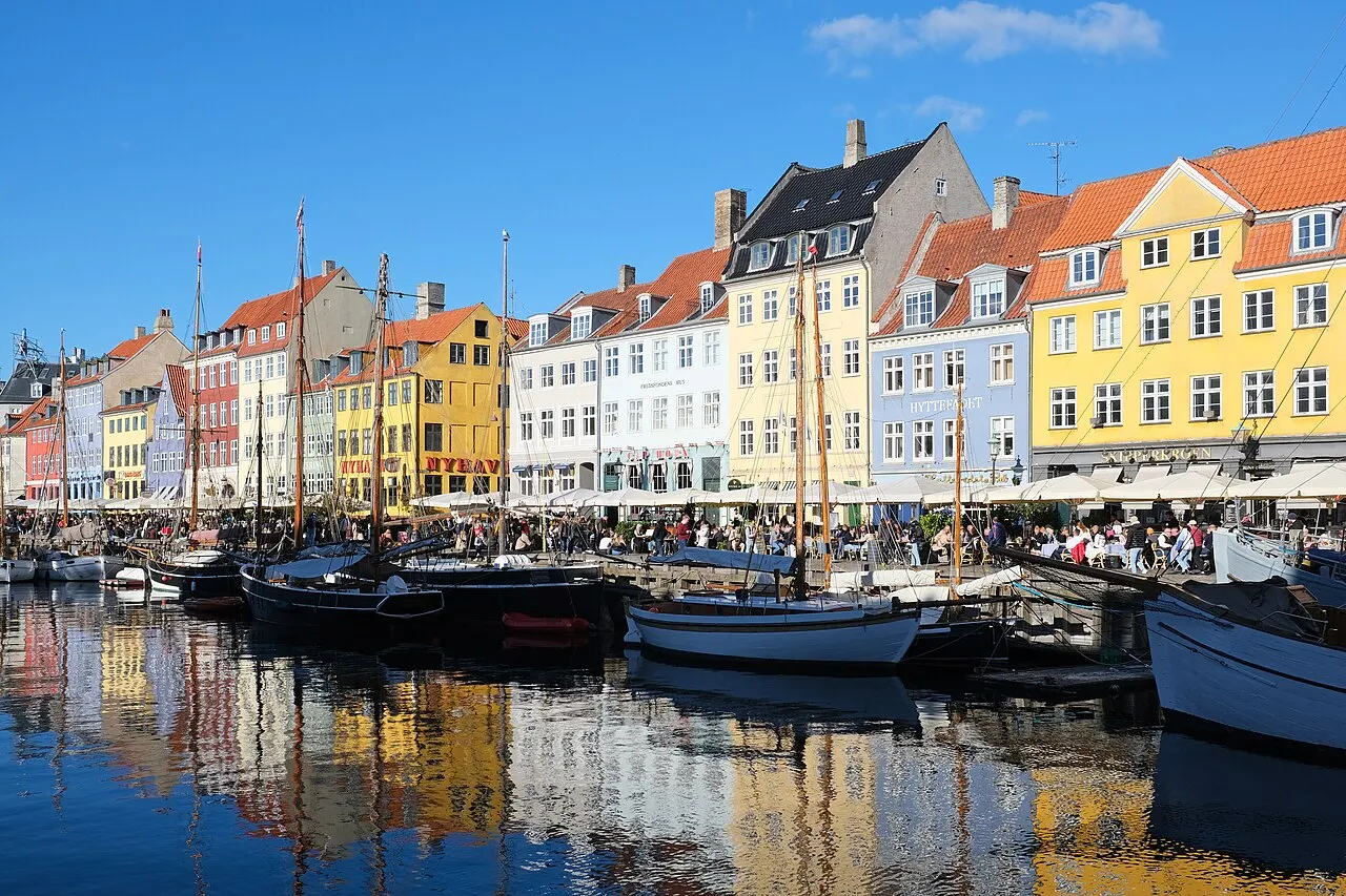 Um canal ladeado por casas estreitas de fachadas coloridas em vermelho, amarelo e branco, com veleiros de mastros altos ancorados na água e pessoas sentadas em mesas ao ar livre na margem, sob céu azul.