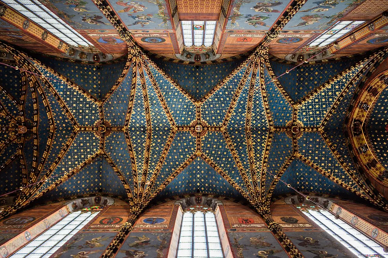 Uma abóbada gótica nervurada, pintada de azul escuro com estrelas douradas, com pinturas religiosas coloridas nos painéis das paredes e vitrais estreitos deixando entrar luz, vista de baixo para cima.